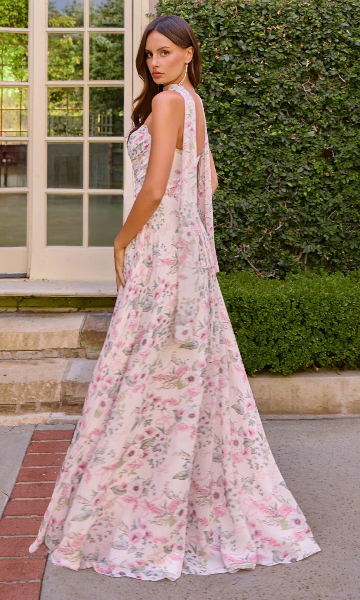 A woman stands outdoors in front of greenery and a windowed door, wearing the Amoris T717 Strapless Long Print Prom Dress by Cinderella Divine—a floral-print gown in pastel colors.